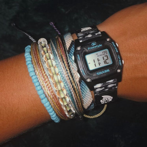 A wrist with colorful beaded bracelets and a Freestyle SHARK MINI CLIP SHARK SCHOOL digital watch with numbers on its display.
