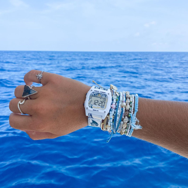 A hand with silver rings, bracelets, and a Freestyle SHARK CLASSIC CLIP OCTOPUS watch held up to the blue ocean sky.