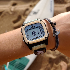 A wrist wearing a Freestyle SHARK CLASSIC CLIP GREEN TEA watch and bracelet, with a sandy beach and surfboard in the background.