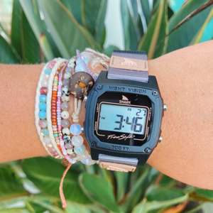 A wrist with colorful beaded bracelets and a Freestyle SHARK CLASSIC LEASH SAHARA displaying numbers, surrounded by green leaves.