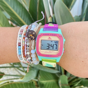 A Freestyle SHARK CLASSIC CLIP SOUR APPLE watch and beaded bracelets on a wrist, with green leaves in the background.