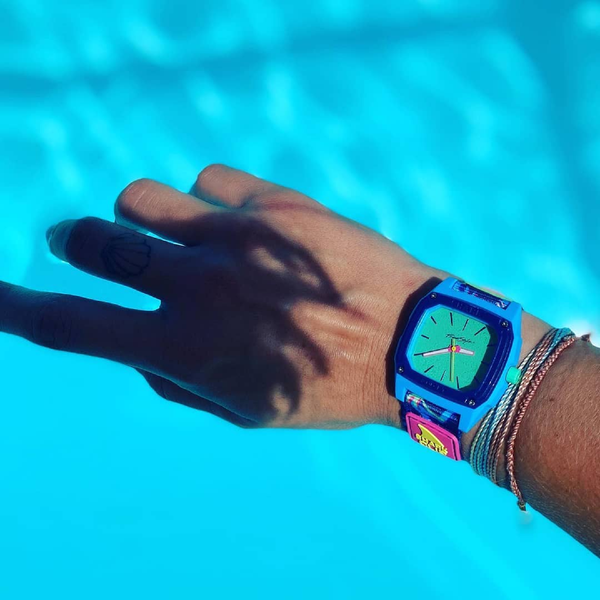 A hand wearing a water-resistant Freestyle SHARK CLASSIC CLIP ANALOG MALIBLU watch and bracelets reaches over a pool.