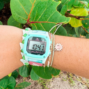 A wrist wearing a Freestyle SHARK CLASSIC CLIP TIE DYE AQUA CLOUD watch, with green leafy plants in the background.