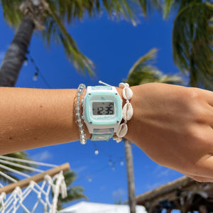A hand with a Freestyle SHARK CLASSIC CLIP TRIPPY TURTLE AQUA watch and shell bracelet is raised beneath bright palms.