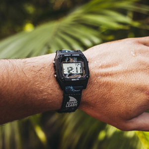 Someone’s wrist wearing a Freestyle SHARK CLASSIC LEASH OCTOPUS BLACK watch, with green palm leaves blurred behind.