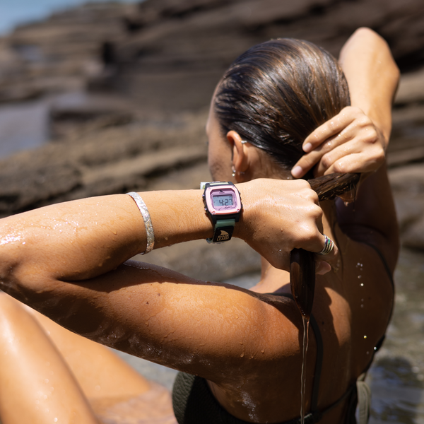 A woman by rocky water squeezes her wet hair, wearing a Freestyle SHARK CLASSIC LEASH ROSE TEA watch on her wrist.