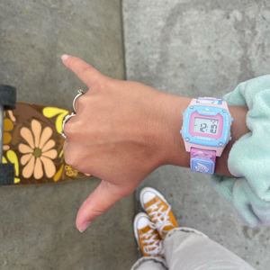 A hand wearing a Freestyle SHARK MINI CLIP TRIPPY TURTLE watch makes a shaka over a floral skateboard and yellow sneakers.