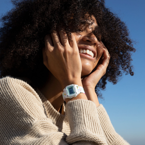 A smiling person with curly hair in a beige sweater wears a Freestyle SHARK MINI CLIP REEF LIFE watch under blue skies.