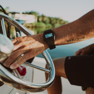 A person with a KAMAʻAINA tattoo steers a boat, wearing a Freestyle SHARK CLASSIC TIDE 600 BLACK (POS) watch.