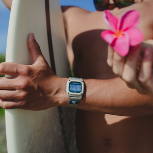 Shirtless surfer with a Freestyle SHARK CLASSIC CLIP COOL SHORE watch holds a pink flower; sky reflects in sunglasses.