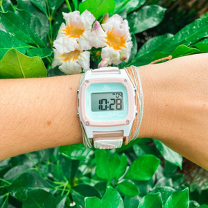 A wrist wears a Freestyle SHARK CLASSIC LEASH BASHFUL watch with numbers displayed, set against green leaves and white flowers in the background.