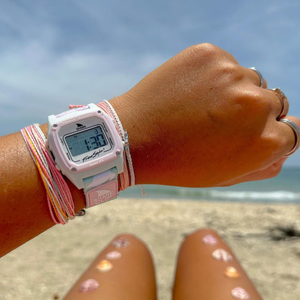 A tan wrist wears the Freestyle SHARK CLASSIC CLIP TWO TONE TURTLE watch and colorful bracelets, with a beach in the background.