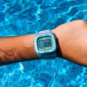 A wrist wearing a Freestyle SHARK CLASSIC CLIP SEA GLASS digital watch held above a pool, with numbers displayed on the screen.