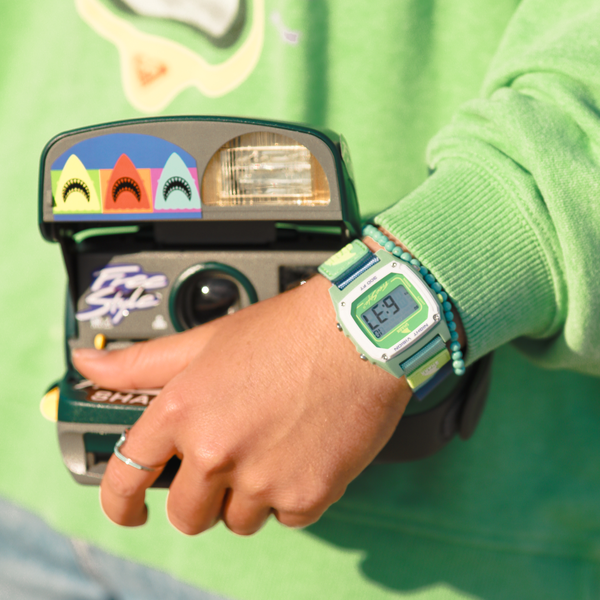 Person in a green outfit holds a retro camera, wearing a matching green Freestyle Caroline Marks Signature Shark Classic Leash watch.