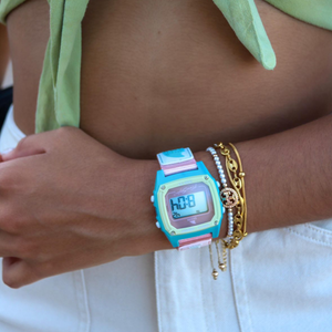 A person wearing a blue Freestyle SHARK CLASSIC LEASH CABO watch and gold bracelets rests their hand on white pants.