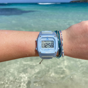 A wrist with a Freestyle SHARK CLASSIC CLIP SKY digital surf watch and bracelets held above clear blue ocean water.