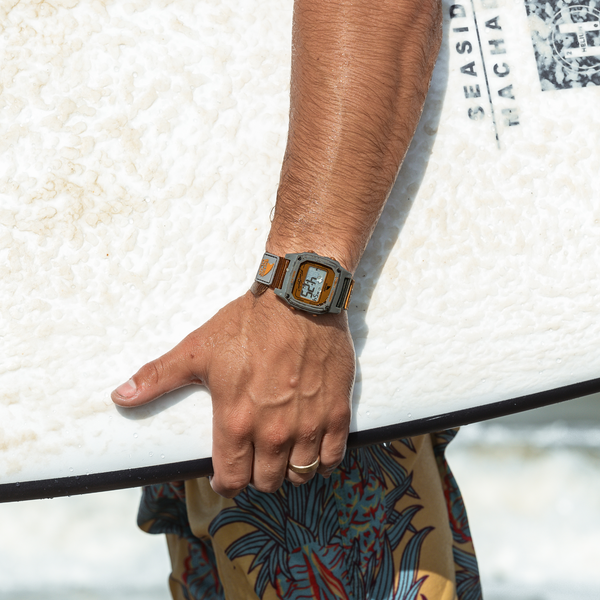 Close-up of a person by the ocean, holding a surfboard and wearing colorful shorts and Freestyles SHARK CLASSIC CLIP MOJAVE watch.