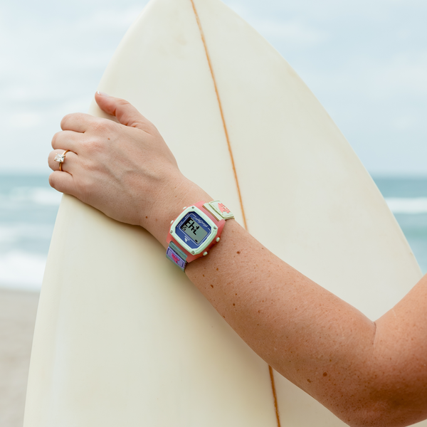 A person wearing a Freestyle SHARK CLASSIC CLIP CORAL BAY watch and a ring holds a surfboard on a cloudy beach.