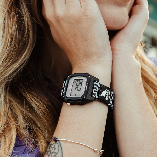 Person outdoors wearing a Freestyle SHARK CLASSIC CLIP ICON watch, with long hair, tattooed arm, and resting chin on hands.