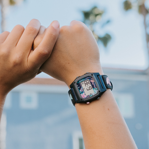 Someone wearing a Freestyle SHARK CLASSIC LEASH MIDNIGHT PALMS watch raises their hand near palm trees and a blue building.