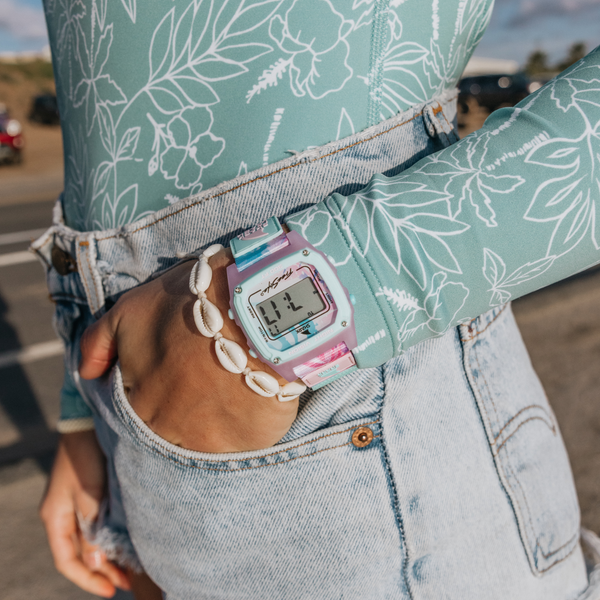 Person wearing a Freestyle Shark Classic Leash Bimini Palms watch, a shell bracelet, a floral long-sleeve shirt, and denim shorts.