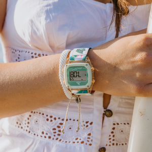 A woman in a white dress wears a Freestyle SHARK CLASSIC CLIP PARADISE PEACH watch and braided bracelets on her left wrist.