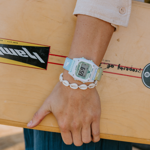 Wearing a Freestyle SHARK CLASSIC LEASH BOHO LIME watch and shell bracelet, a person stands holding a skateboard by their side.