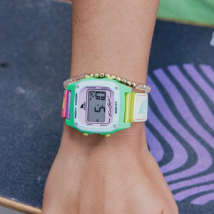 Someone wearing a Freestyle SHARK CLASSIC LEASH WATERMELON watch stands next to a purple-striped skateboard.