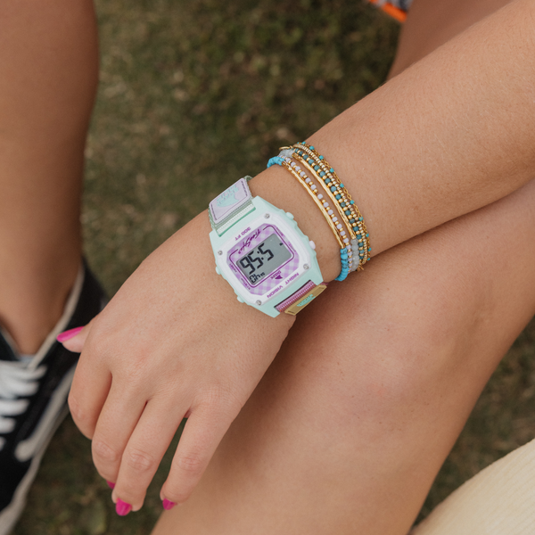 A person outdoors wearing a Freestyle SHARK CLASSIC CLIP watch in Gingham Pink and colorful beaded bracelets, with their hand resting on their knee.