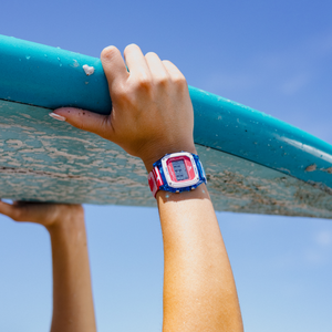 A person wearing a Freestyle SHARK CLASSIC CLIP FINS & TAILS watch holds up a blue surfboard beneath a clear sky.