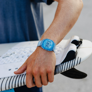 A person wearing a Freestyle SHARK CLASSIC CLIP BLUEBERRY watch holds a white surfboard featuring black stripes.