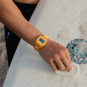 Wearing a Freestyle SHARK CLASSIC CLIP TANGERINE, someone waxes a white surfboard on the sand.