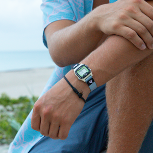 Close-up of a wrist wearing the Freestyle SHARK MINI CLIP ACAI watch and a black string bracelet near the beach.