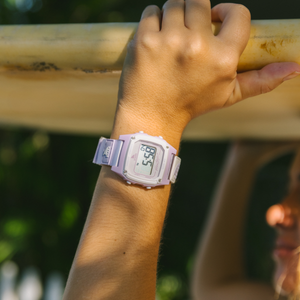 Wearing a Freestyle SHARK CLASSIC LEASH LILAC SPLASH, a person holds up a surfboard outdoors under bright natural light.