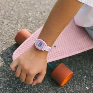 Someone wearing a Freestyle SHARK MINI LEASH PINK CRUSH watch rests their hand on a pink skateboard with orange wheels.