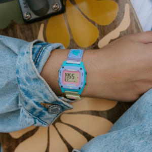 Wearing a denim jacket, someone shows off a Freestyle SHARK MINI CLIP LAVENDER TEA watch while standing on a floral skateboard.