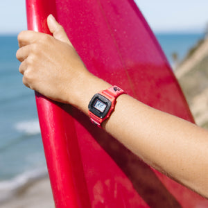 Person holding a red surfboard with a digital watch on wrist, beach background