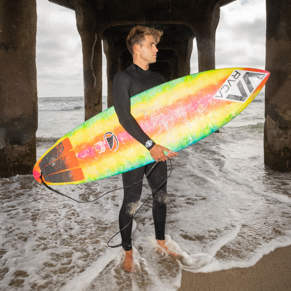 A surfer in a wetsuit stands beneath a pier, holding a board featuring a Freestyle Fin Waterproof Decal by Freestyle.