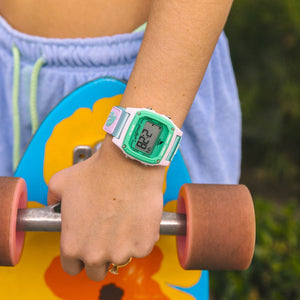 Wearing a Freestyle SHARK CLASSIC CLIP PURE AQUA watch, someone holds a skateboard featuring orange wheels and yellow flowers.
