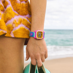 A person in a colorful towel on the beach holds shoes and wears a Freestyle SHARK CLASSIC LEASH NEON SEA watch.