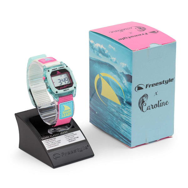 A colorful digital wristwatch, the CAROLINE MARKS SIGNATURE SHARK CLASSIC LEASH CLEAR BLUE SKY, is displayed on a black stand beside a matching box. Featuring a turquoise and pink design, the box showcases a wave graphic and the Freestyle logo, celebrating their collaboration with Caroline Marks.