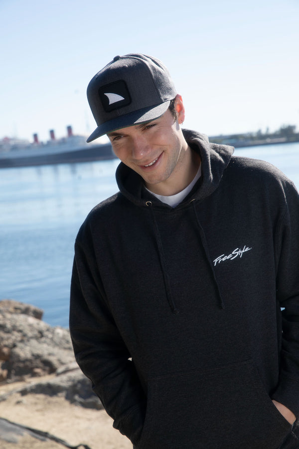 Smiling man in a black hoodie and Freestyle Shark Fin Snapback Hat Heather Grey stands by water with ship and rocks.