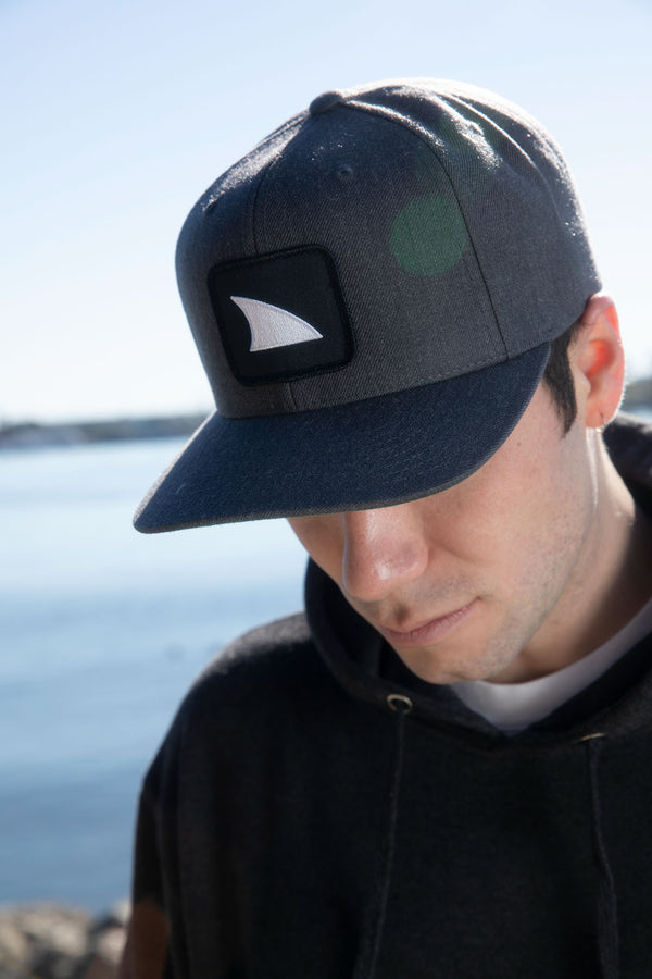 Man wearing the Freestyle Shark Fin Snapback Hat in Heather Grey, looking down by the water on a sunny day.