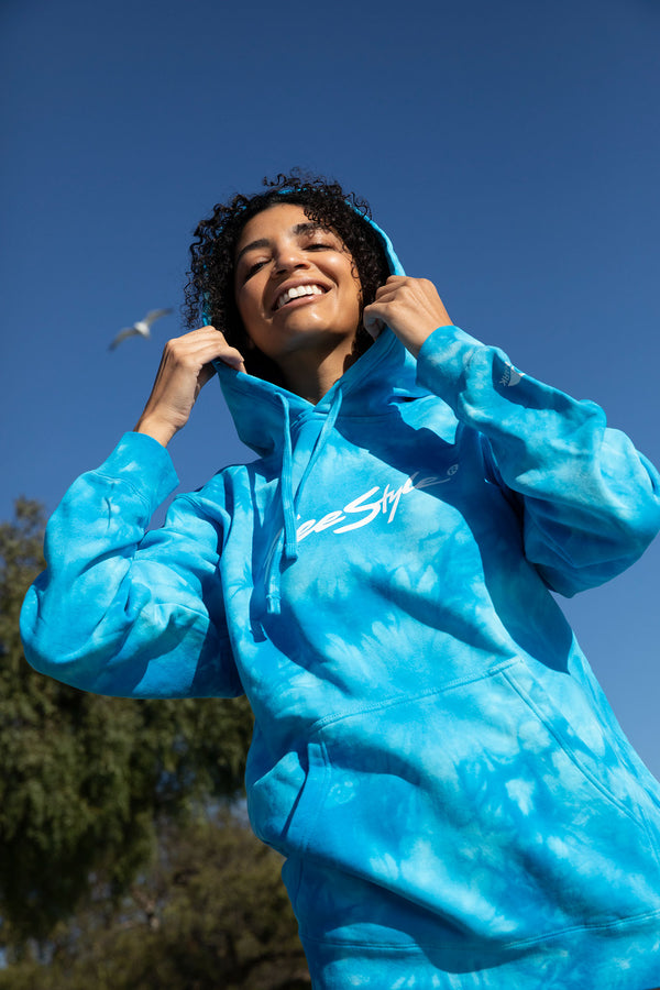 Person smiling outdoors in a Freestyle Unisex Aqua Dream Hoodie beneath a clear blue sky.
