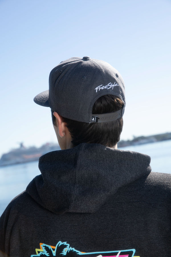 Person in a dark hoodie and Freestyle Shark Fin Snapback Hat Heather Grey stands by water, ship blurry in background.