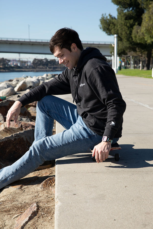 Person sitting on a ledge by the water in a Freestyle Unisex 40th Neon-versary Hoodie and jeans, with a skateboard beside them.