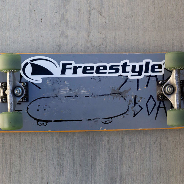 A skateboard with green wheels and a Freestyle/Fin Large Waterproof Decal resting on concrete.